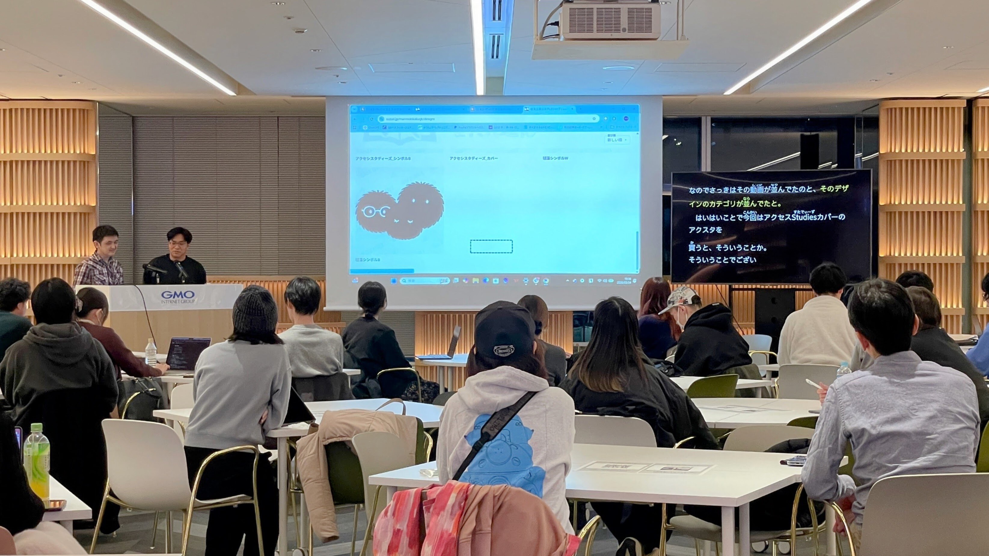 ウォークスルー中の会場の様子。参加者が着席して見守るなか、登壇者2名がスクリーン脇に立っている。メインスクリーンにはSUZURIのページが映し出され、サブモニターにはUDトークの字幕が表示されている